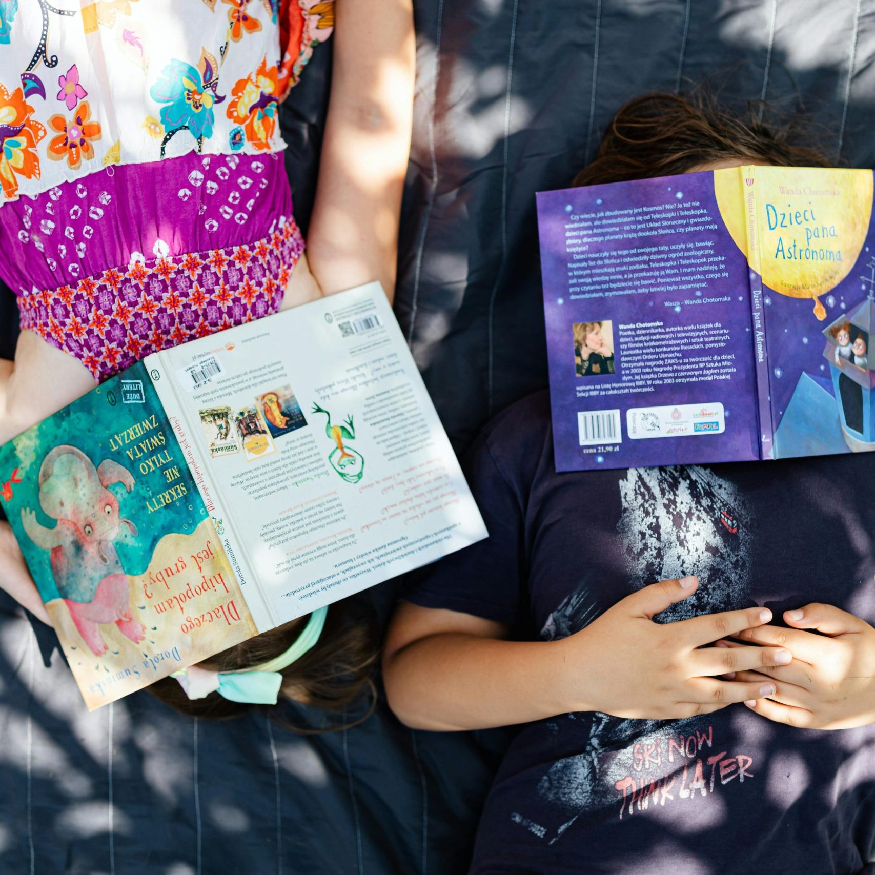 Two children lie outdoors on grass, faces covered by books, enjoying a sunny day.