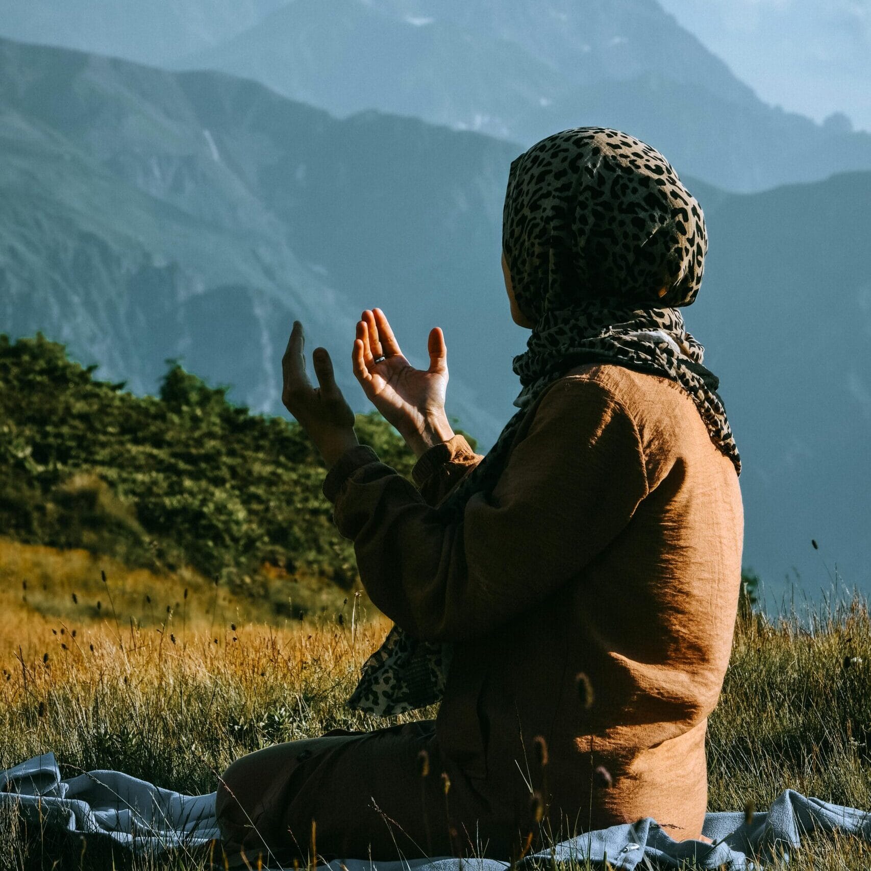 A woman in a headscarf prays outdoors with mountains in the background.