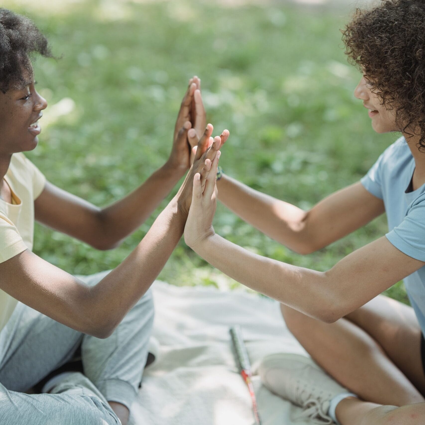 Two girls enjoy hand games outdoors on a blanket during a sunny day picnic.