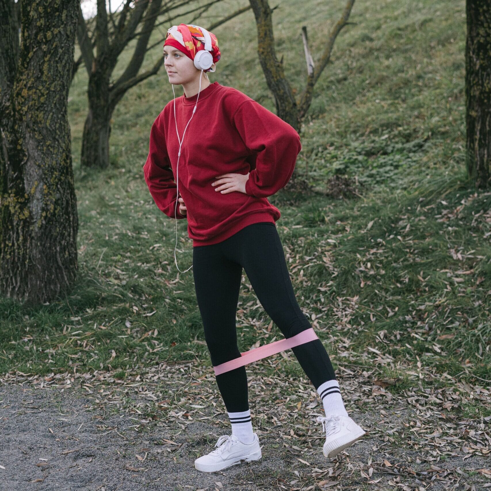 A woman working out in a park wearing headphones and a headscarf, using a resistance band.