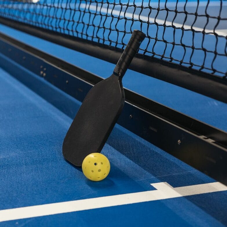 Pickleball paddle resting against net with yellow ball on blue court