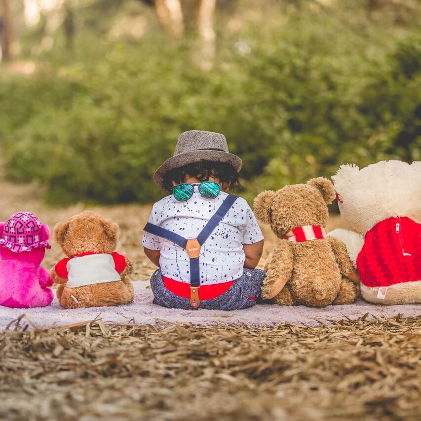 A toddler sitting outdoors with plush toys, enjoying the scenic nature in a park.