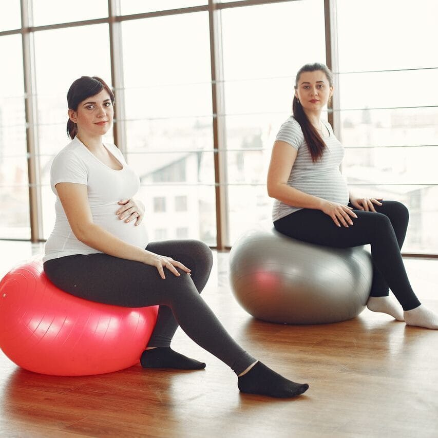 Positive pregnant ladies sitting on fit balls during prenatal training in spacious light gym at daytime and smiling while looking at camera